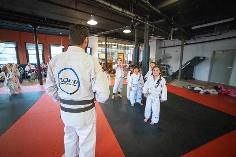 Martial arts class in a gym, instructor facing students in white gis on red and black mats.