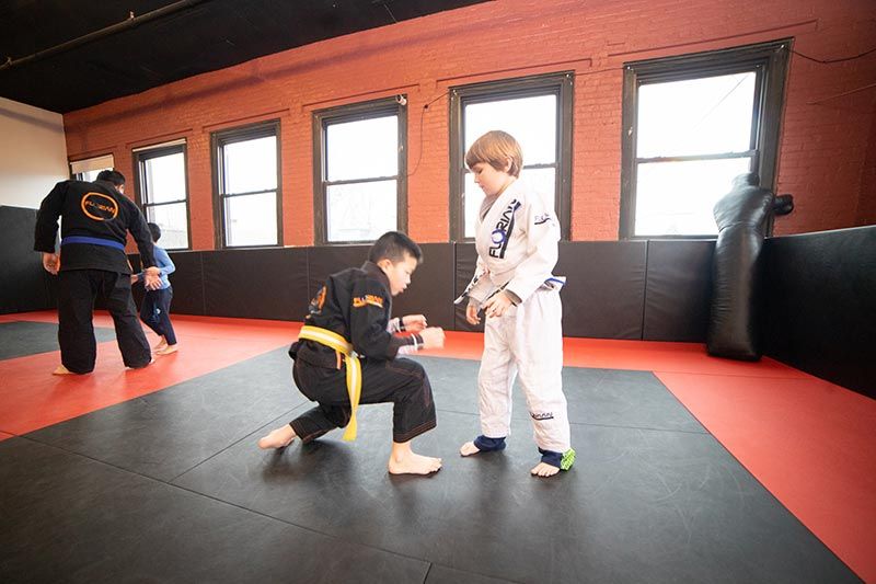 Children in martial arts uniforms practicing grappling on red and black mats in a training room