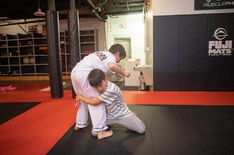 Two martial artists practice a grappling hold on mats in a gym with boxing equipment.