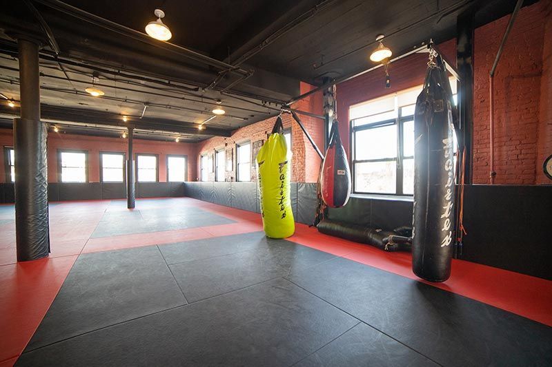 Empty martial arts gym with red-and-black mats, hanging punching bags, and brick walls.