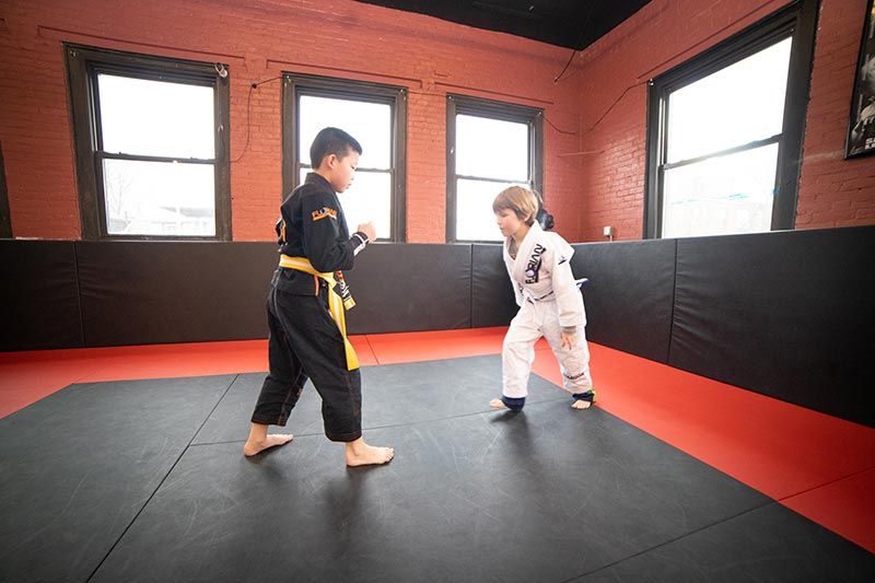 Two children in martial arts uniforms spar on a padded gym floor in a bright room with windows.
