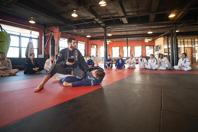 Brazilian jiu-jitsu class training on mats in a gym, instructor demonstrating a hold while students watch.
