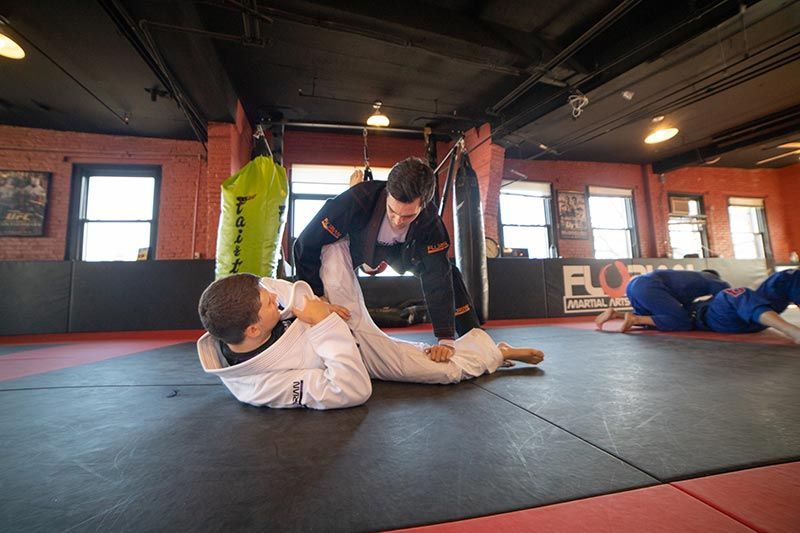 People grappling on mats in a martial arts gym, with one person pinning another.