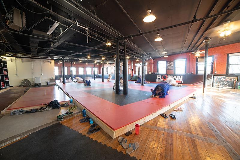 Large martial arts studio with red mats, black columns, and hanging lights; gear scattered on the floor.