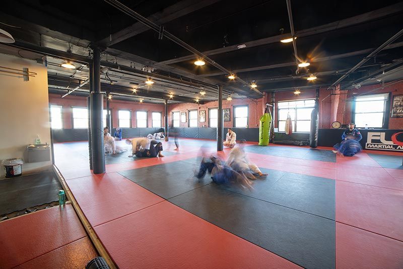 Indoor martial arts gym with people grappling on red-and-black mats near large windows