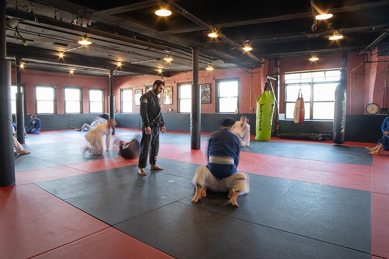 People training on mats in a martial arts studio with large windows and overhead lights.