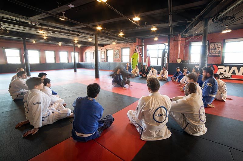 Martial arts class seated on mats in a red and black training gym, watching an instructor.