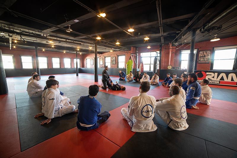 People sitting on mats in a martial arts studio, facing an instructor on red-and-black flooring.