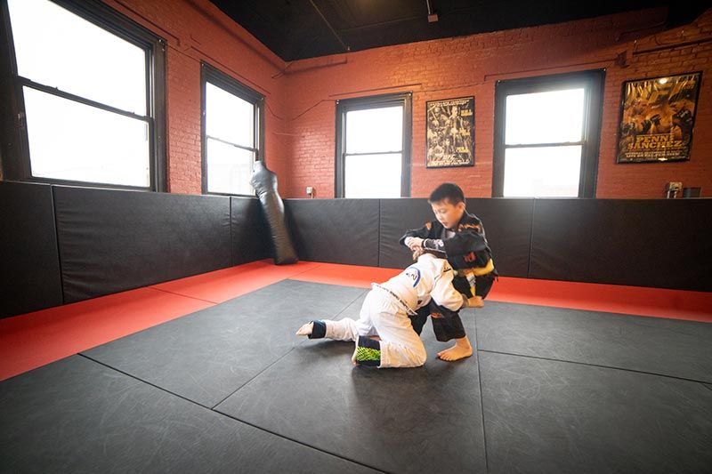 Two people grappling on a red-and-black mat in a martial arts gym with brick walls and windows