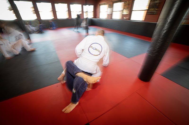 Person seated on red and black martial arts mats in a dojo, with others blurred in the background