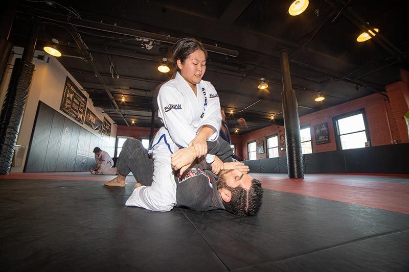 Two martial artists grappling on mats in a dojo, one in a white gi pinning the other.