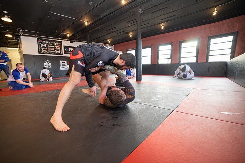 Two men grappling on mats in a martial arts gym, with others training in the background.