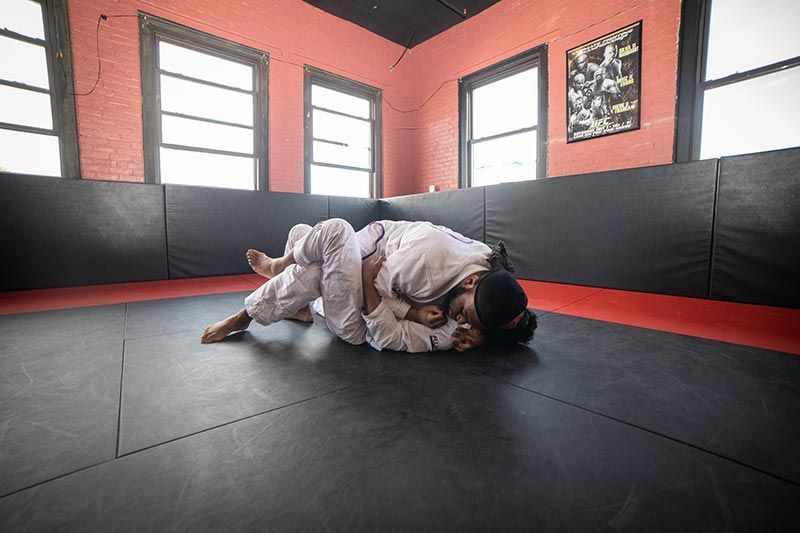 Two people grappling on a padded martial arts mat in a gym with red walls and windows