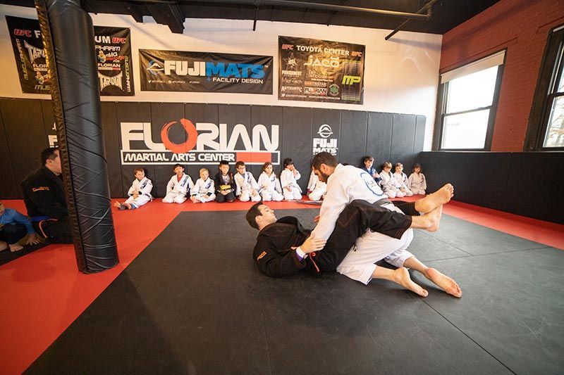People practicing grappling on black mats in a martial arts gym with spectators watching