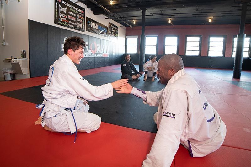Two people in white gis practice a handshake drill on red mats in a martial arts studio.