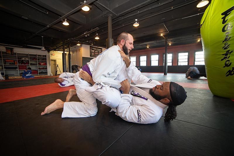 Two martial artists practicing grappling on mats in a gym, one pinning the other.
