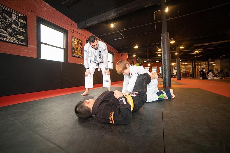 Two people practicing grappling on mats while a coach watches in a gym.