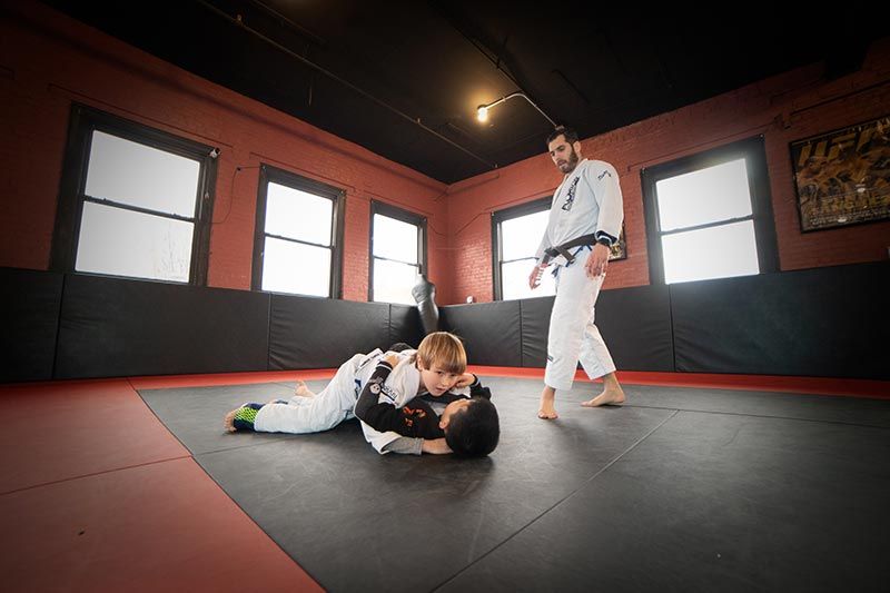 Two martial artists grappling on mats in a dojo, with a standing instructor in the background.