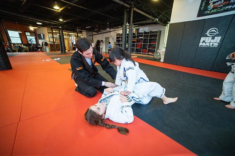Martial arts class on orange mats, instructor demonstrating a grappling hold with two students.