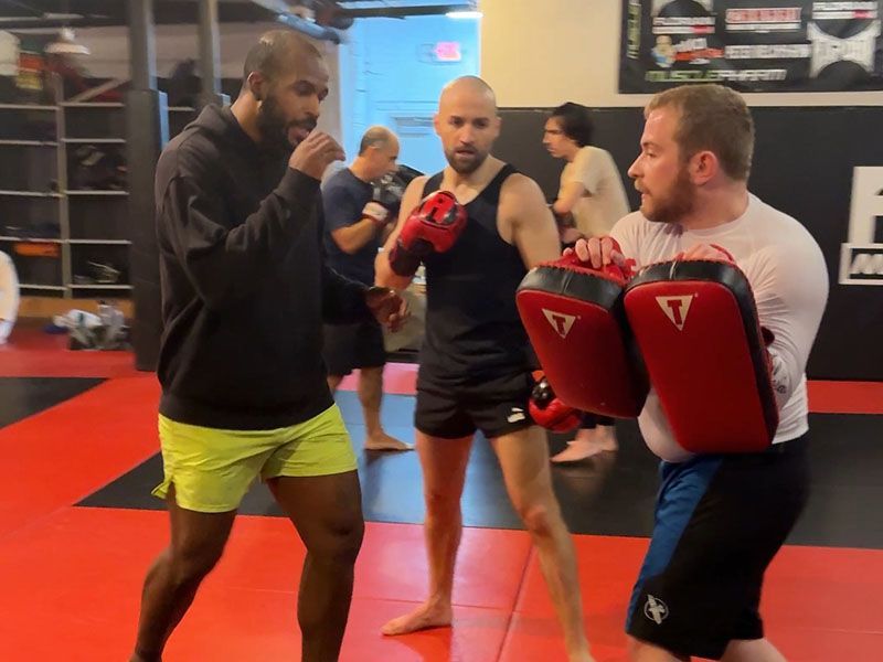 Three men in a martial arts gym practice pad drills on red mats.