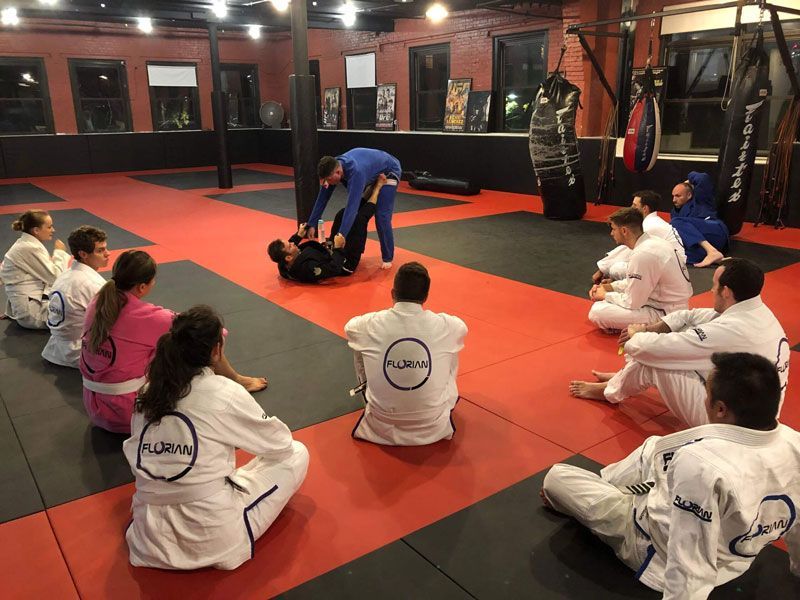 Martial arts class on red mats, students seated around two people demonstrating a grappling technique.
