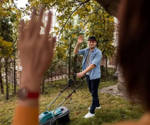Two people waving in a wooded park, one in the foreground and one near a lawn mower.