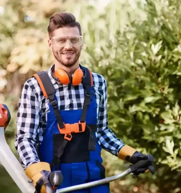 Person in blue overalls holding a string trimmer in a sunny garden, with orange safety gear.