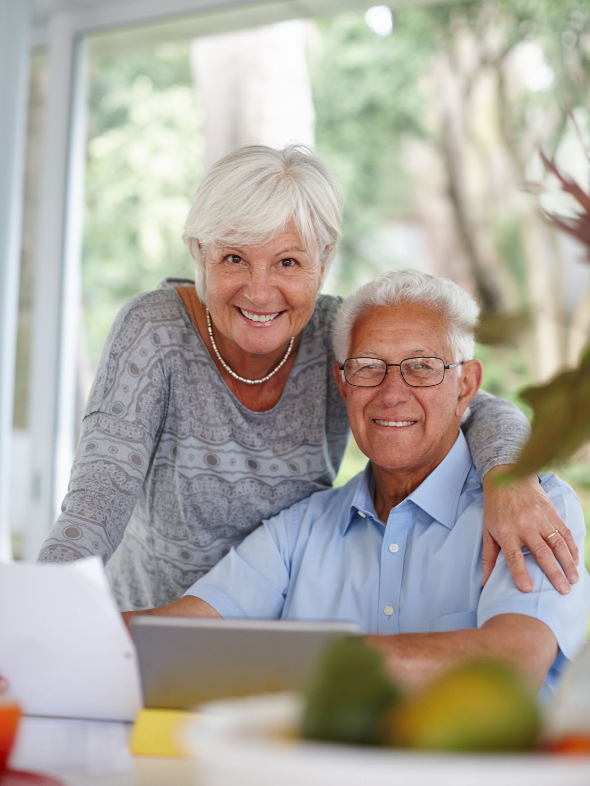 Smiling elderly couple indoors; woman has arm around man; they are near a table with a laptop.
