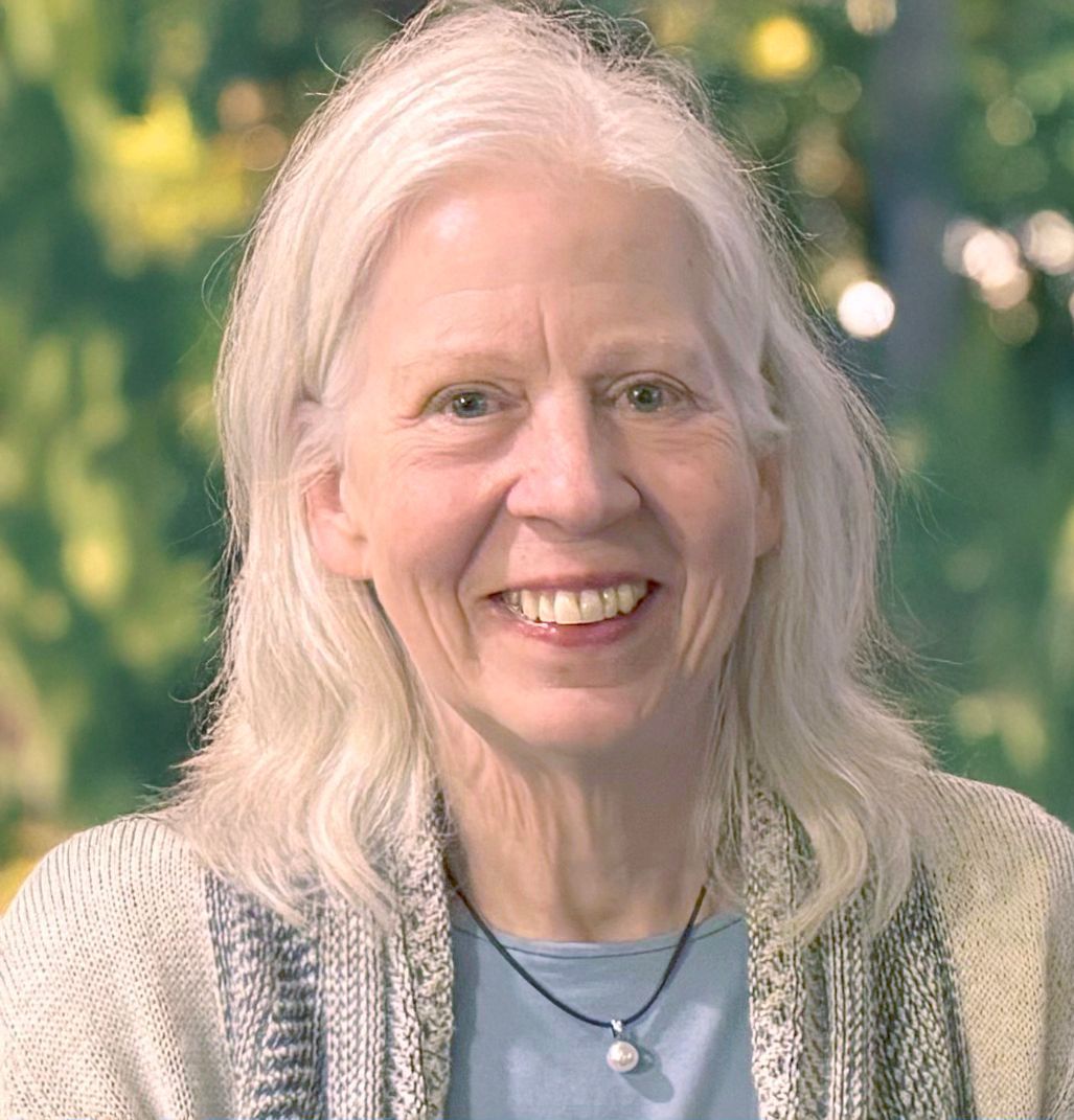 Woman with long white hair smiles, wearing a light sweater and blue shirt; blurry outdoor background.