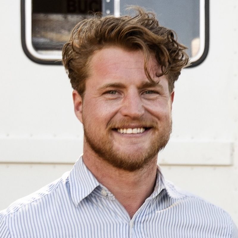 Man with curly red hair and beard smiling, wearing a blue striped shirt in front of a white truck.