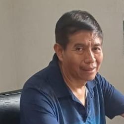 Man with dark hair in a blue shirt, smiling. Indoors, against a plain wall.