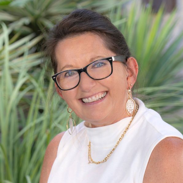 Smiling woman with glasses and gold jewelry in front of greenery.