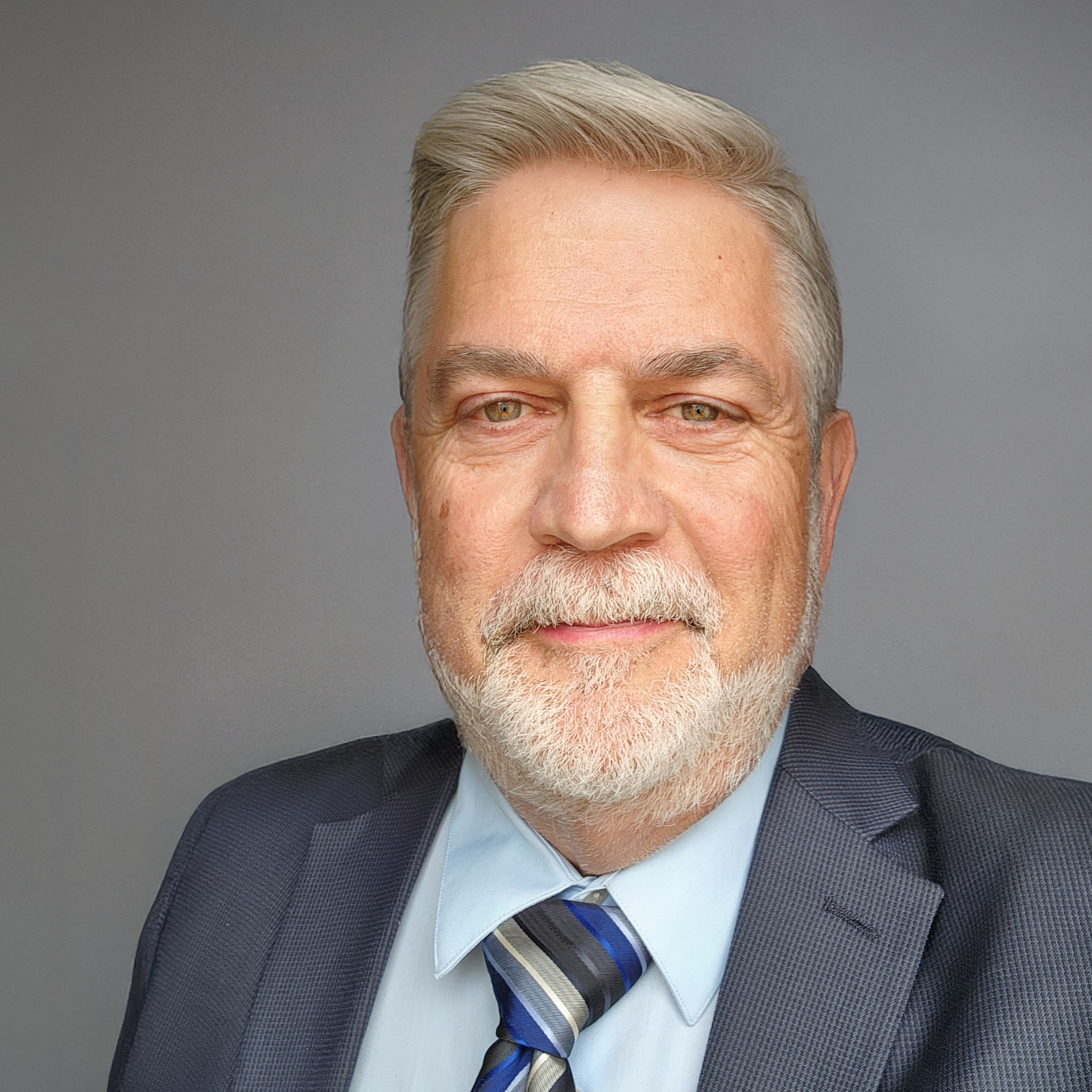 Man with graying hair and beard, wearing a suit and tie, against a gray background.