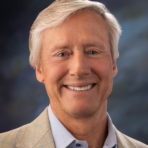 Smiling man with gray hair, wearing a light-colored blazer, blue shirt, against a blurred background.
