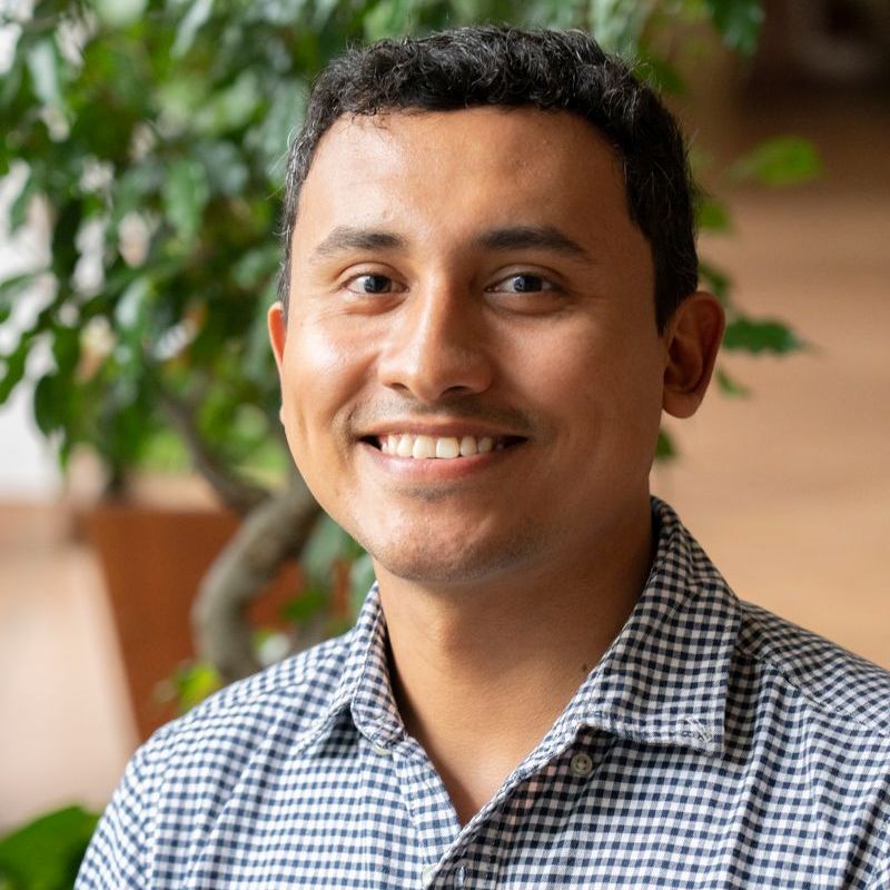 Smiling man in blue checkered shirt, indoors, plants in background.