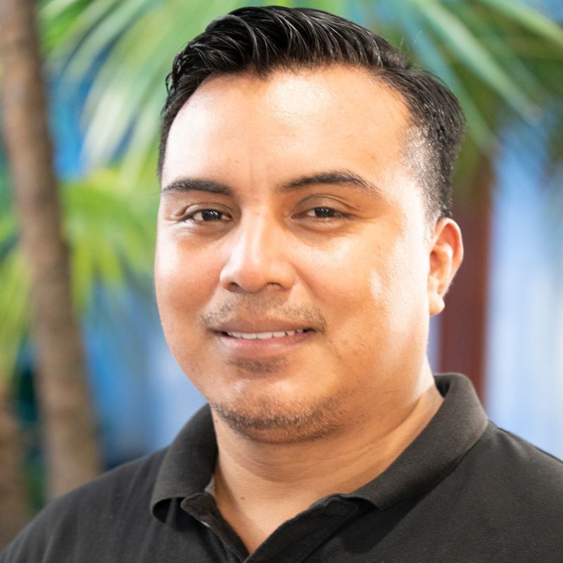Man with dark hair and black shirt smiles, palm tree in background.