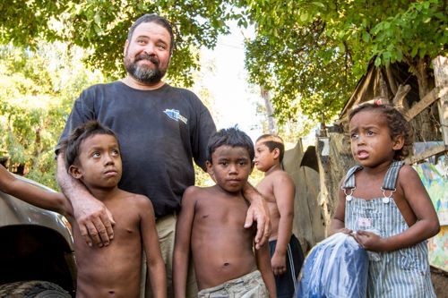 Man with beard and shirtless children outdoors; smiles, in a sunlit, impoverished area.