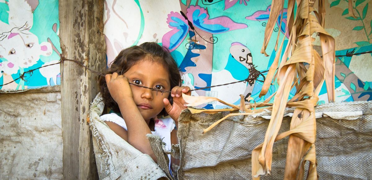 A young girl with glasses, sitting in a shelter, looks directly at the camera.