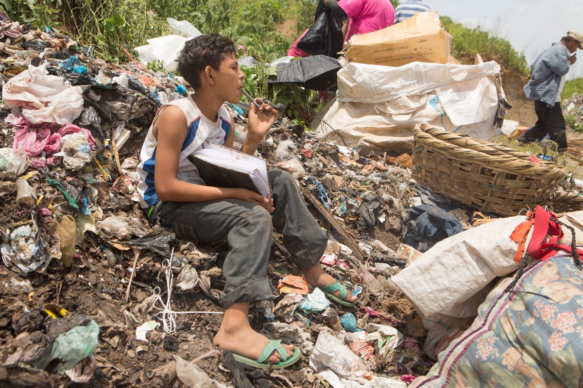 Boy sits on a trash heap, holding a book and a small item, while others sort through the garbage.