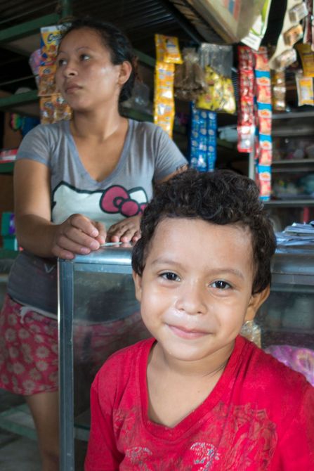 A young boy smiles, wearing a red shirt, standing in front of a woman at a store counter.
