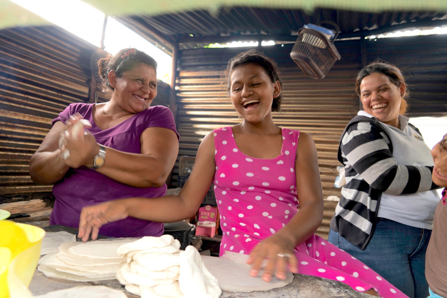 Three smiling women make tortillas outdoors. One presses dough.