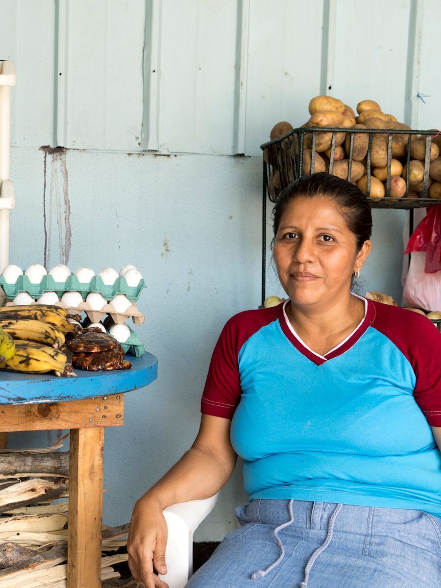 Woman at a market stall; eggs, bananas, potatoes visible. Blue and red shirt, blue skirt.