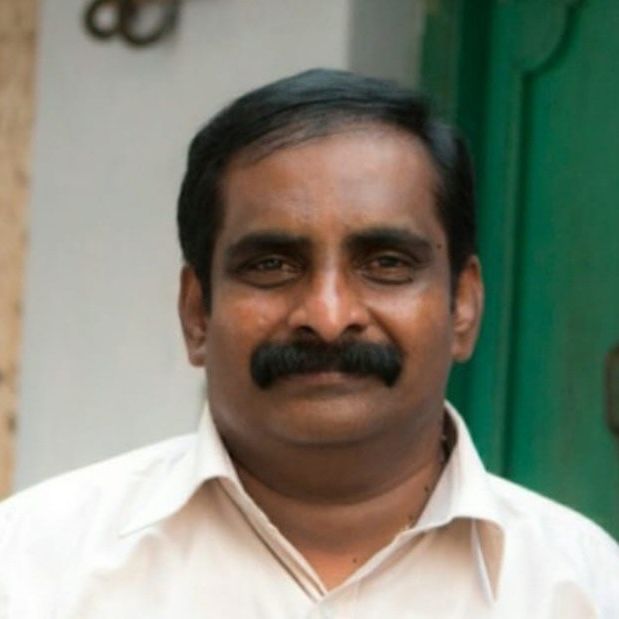 Man with dark hair and mustache smiles, wearing a light shirt, against a blurred outdoor background.