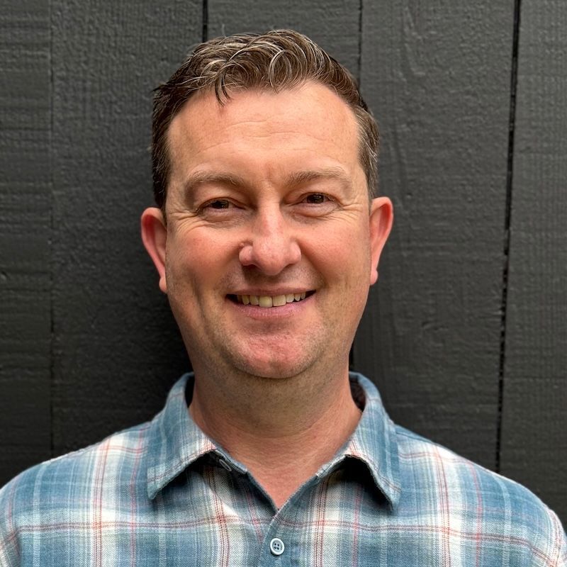 Man with fair skin and brown hair smiles in a plaid shirt against a dark wood wall.
