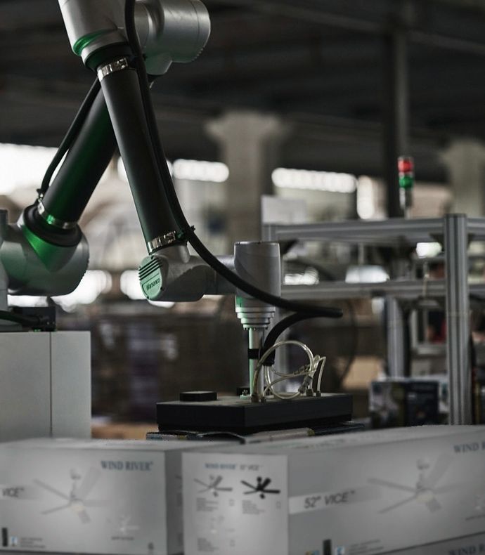 A robotic arm in a factory places a ceiling fan part onto a box. The arm is metallic green and black.