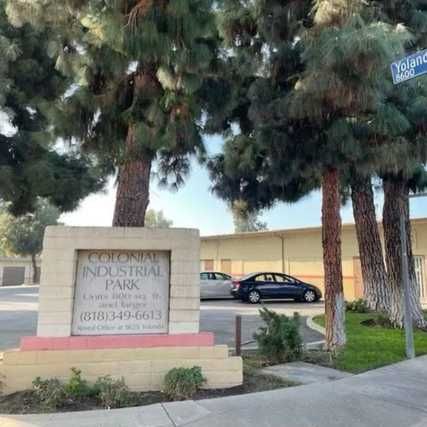 A photo of tall pine trees standing beside a street corner with a sign that reads “Colonial Industrial Park”