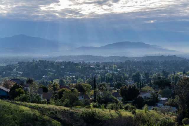 A wide view of a valley town with houses, trees, and hills under a cloudy sky with soft sun rays shining through.