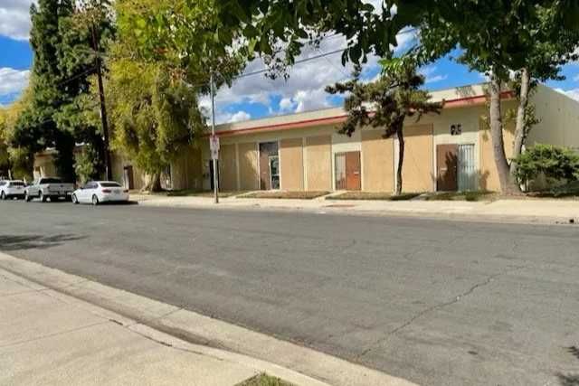 A quiet street lined with parked cars faces a long, low building with beige walls and a red trim.