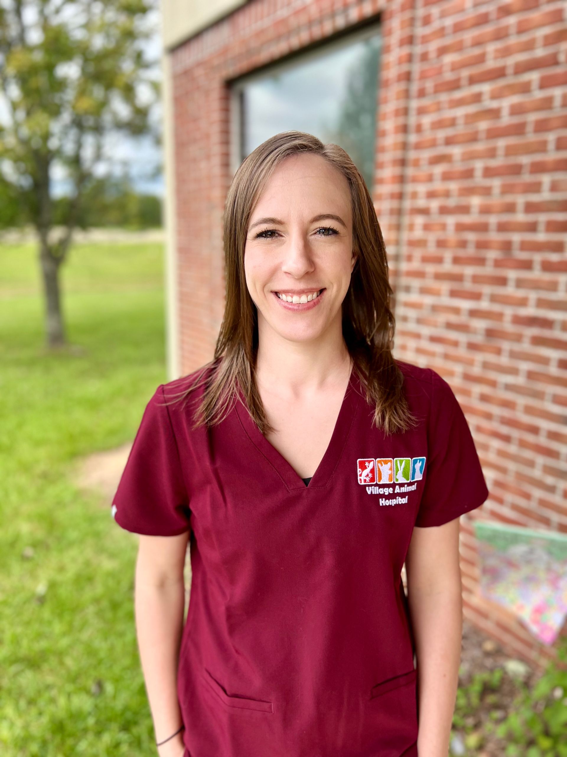 A woman in a maroon scrub top is smiling in front of a brick building.