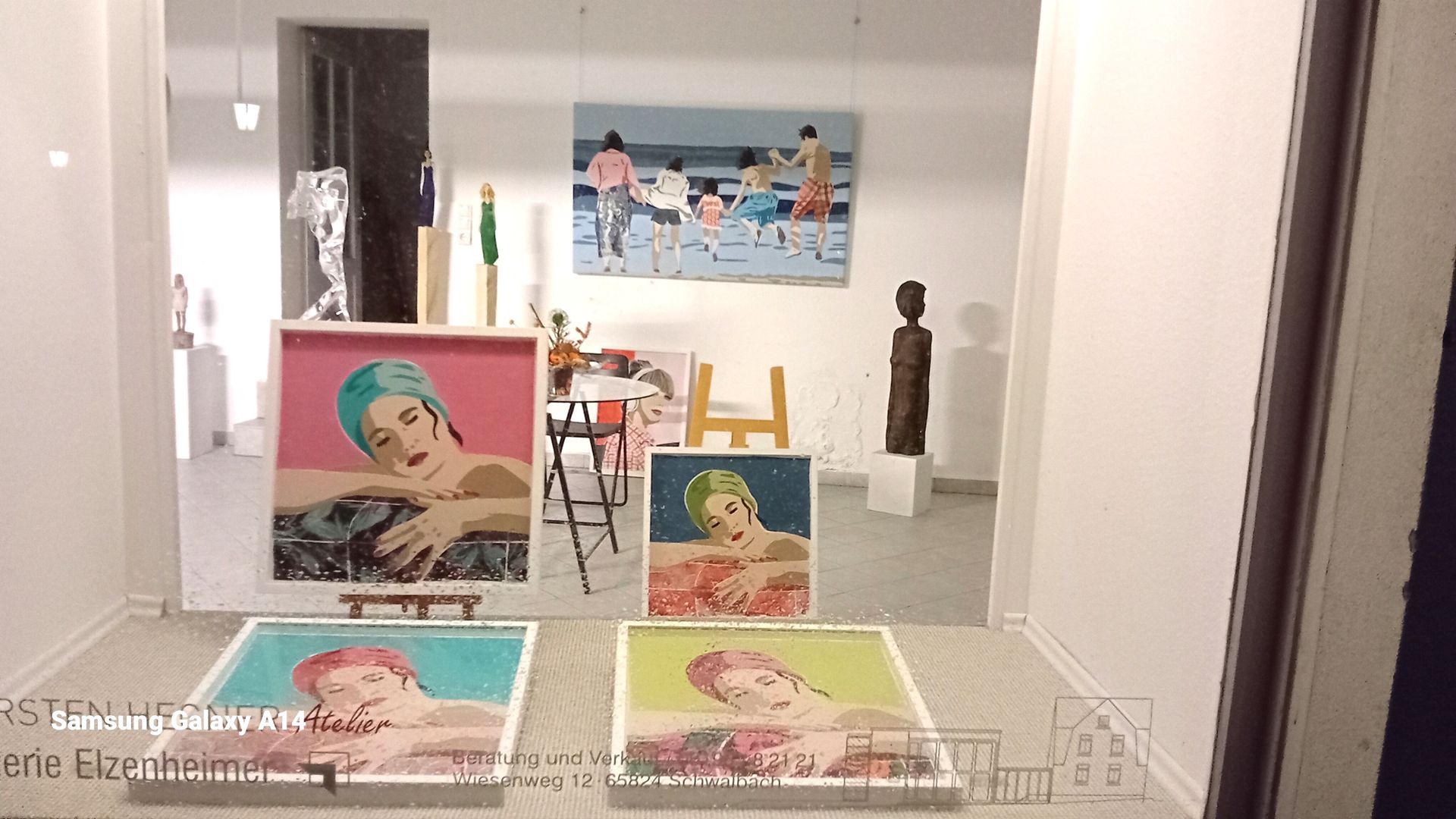 Schaufenster einer Kunstgalerie mit Gemälden von Frauen mit Badekappen und einem Gemälde mit Menschen an einem Strand.